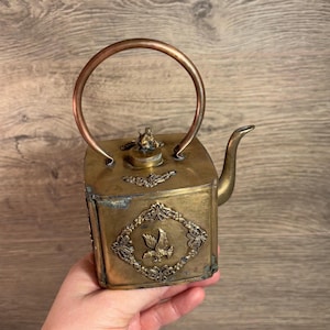 Peut inclure: Une petite théière carrée en laiton avec un design orné. La théière a une poignée en cuivre et un bec verseur. Le design présente un oiseau en vol et des motifs floraux.