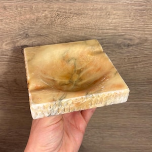 Puede incluir: Un cenicero cuadrado de mármol marrón claro con una depresión circular tallada en el centro. El cenicero tiene un borde texturizado y tallado.