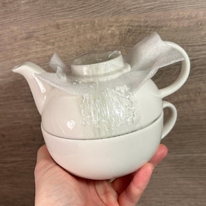 Puede incluir: Juego de tetera y taza de cerámica blanca. La tetera tiene un cuerpo redondo y un pico. La taza es de forma de cuenco grande. El juego está envuelto en un embalaje protector.