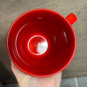Peut inclure: Une tasse en c&eacute;ramique rouge vif avec une forme de bol arrondie et une petite anse. L'int&eacute;rieur de la tasse est rouge brillant, refl&eacute;tant la lumi&egrave;re. La tasse est tenue dans une main, avec une surface en bois en arri&egrave;re-plan.
