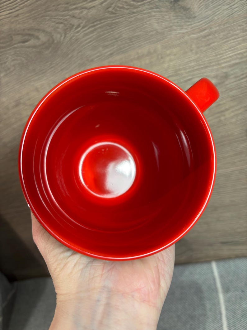 Peut inclure: Une tasse en c&eacute;ramique rouge vif avec une forme de bol arrondie et une petite anse. L'int&eacute;rieur de la tasse est rouge brillant, refl&eacute;tant la lumi&egrave;re. La tasse est tenue dans une main, avec une surface en bois en arri&egrave;re-plan.