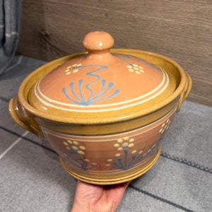 Könnte beinhalten: Ein runder Keramik-Schmortopf mit passendem Deckel, der eine gesprenkelte senfgelbe Basis und ein pfirsichfarbenes Mittelband aufweist. Die Oberfläche ist mit handgemalten blauen Blumenmotiven und kleinen weißen Punkten in kreisförmigen Mustern verziert.
