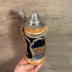 May include: A ceramic beer stein with a pewter lid and a scene of a German town with the word "Heidelberg" on the bottom. The stein is decorated with blue, red, green, and yellow colors.