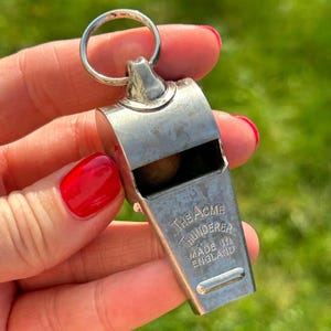 Puede incluir: Silbato Acme Thunderer plateado con anilla. El texto "THE ACME THUNDERER MADE IN ENGLAND" está grabado. El silbato se muestra sobre un fondo verde borroso.