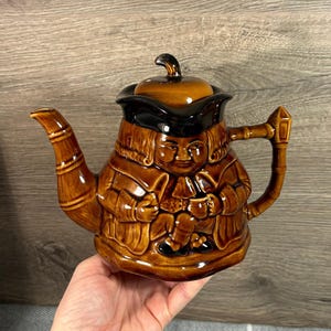 Puede incluir: Una tetera de cerámica marrón con un diseño de figura. La tetera presenta a un hombre con sombrero y abrigo, con asa y boquilla. La tapa es marrón y negra. Es un artículo decorativo.