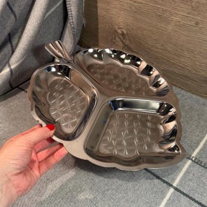 Puede incluir: Una fuente plateada en forma de hoja con tres compartimentos. La fuente tiene un interior texturizado con un patrón de diamantes y un borde festoneado. La fuente es sostenida por una mano con esmalte de uñas rojo.