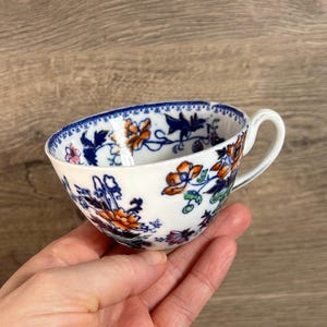 Puede incluir: Una taza de té blanca con un patrón floral azul y naranja. La taza tiene un asa y está decorada con un borde azul y blanco.