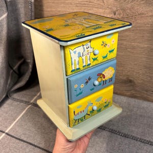 Puede incluir: Una caja de madera con tres cajones, cada uno con un diseño de animal pintado a mano diferente. El cajón superior tiene un fondo amarillo con un gato, el cajón del medio tiene un fondo azul con una oveja y el cajón inferior tiene un fondo amarillo con una vaca. La caja está pintada en un esquema de colores azul claro y amarillo.