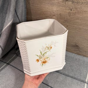 May include: White ceramic planter with a textured basketweave design. The planter features a floral design with yellow and white flowers. The rim is trimmed with a gold accent. The planter is square with an octagonal base.