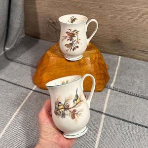 May include: Two white ceramic mugs with bird illustrations. One mug is on a wooden base, and the other is held by a person. The mugs have a blue trim and curved handles. The bird illustrations feature various colorful birds on branches.