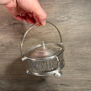 Può includere: Una zuccheriera vintage color argento con coperchio e manico. La ciotola ha un design in vetro tagliato trasparente e poggia su tre piedini. Il manico si incurva sopra la parte superiore e il coperchio ha un piccolo ornamento decorativo.