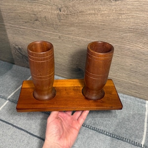 May include: A wooden desk organizer with two cylindrical holders and a rectangular base. The organizer is made of a medium-toned wood with visible grain and is designed to hold pens, pencils, or other small items. The base has a smooth, polished finish.