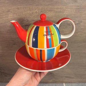May include: A colorful striped teapot and cup set with a red saucer. The teapot and cup are hand-painted with a variety of colors, including red, orange, yellow, green, blue, and purple. The teapot has a red lid and a white label that reads "Whittard of Chelsea Handpainted".