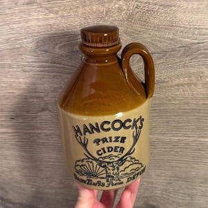Könnte beinhalten: Ein brauner Keramikkrug mit Henkel und Verschluss. Der Krug trägt die Aufschrift "HANCOCK'S PRIZE CIDER" mit einer Hirschillustration. Der Krug wird wahrscheinlich zur Aufbewahrung von Apfelwein oder anderen Getränken verwendet.