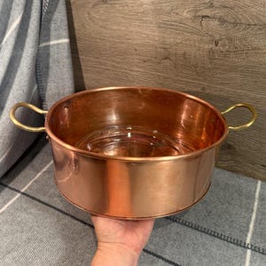 Puede incluir: Un cuenco de color cobre con dos asas de latón. El cuenco tiene forma redondeada y un acabado brillante. El interior del cuenco es visible y las asas son curvas. El cuenco probablemente se utiliza para cocinar o servir.