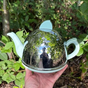 Könnte beinhalten: Eine polierte verchromte Teekanne mit hellblauem Ausguss, Griff und Deckel. Die Teekanne reflektiert die umliegende Vegetation. Die Teekanne steht auf einem hellblauen Sockel.