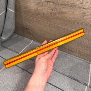 May include: A vintage yellow and red slide rule. The slide rule is rectangular, with a yellow body and red stripes. The slide rule is approximately 12 inches long.