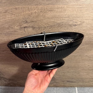 May include: An oval-shaped black pedestal bowl featuring a fluted exterior design. Inside the bowl, there is a white metal grid insert with visible rust spots, held in place by two small metal hooks attached to the rim of the black container.