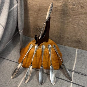 Puede incluir: Conjunto de siete cuchillos de mantequilla de plata con mangos de madera, expuestos en un soporte de madera. Los cuchillos están dispuestos en forma de abanico, con los mangos apuntando hacia el centro del soporte.