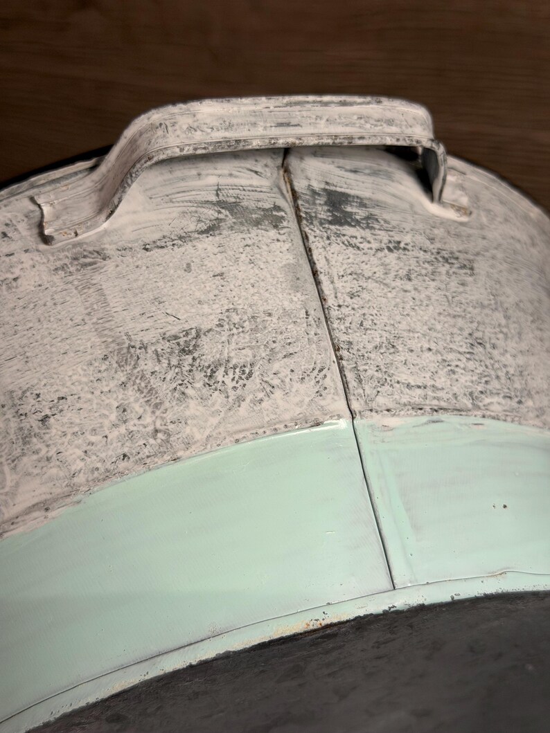 Op de afbeelding: Een metalen emmer met een gebogen handvat en een tweekleurige afwerking. Het bovenste gedeelte heeft een verweerde witte verflaag over een donkere basis, terwijl het onderste gedeelte is geschilderd in een licht mintgroene kleur met een zichtbare naad.
