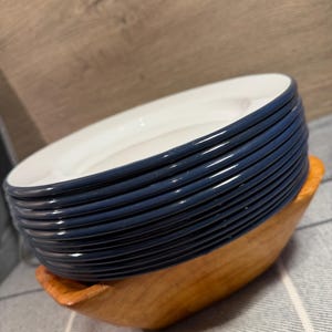 Pode incluir: Uma pilha de dez pratos redondos com centros brancos e bordas azul-escuras, descansando dentro de uma tigela de madeira castanho-clara. Os pratos est&atilde;o dispostos numa pilha vertical organizada, destacando o contraste entre as superf&iacute;cies brancas.
