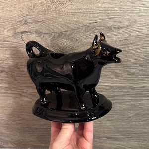Puede incluir: Una cremera de cerámica negra con forma de vaca, con detalles dorados en los cuernos y las orejas. La cremera es brillante y se apoya sobre una base redonda. Un artículo de cocina decorativo.