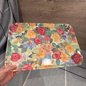 Könnte beinhalten: Rechteckiges Serviertablett mit floralem Muster. Das Tablett hat einen cremefarbenen Hintergrund mit einer lebendigen Anordnung aus roten, rosa, gelben und blauen Blumen und grünen Blättern. Das Tablett besteht aus glänzendem Material.