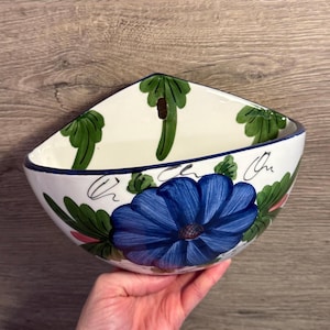 May include: A white ceramic bowl with a blue floral design. The bowl has a triangular shape with a blue rim and is decorated with green leaves and a large blue flower. The bowl is held in a person's hand.