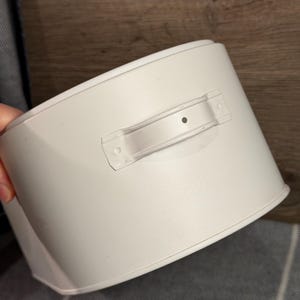 May include: A matte white cylindrical metal container featuring a single horizontal handle attached to the side. The handle is secured with two visible rivets and has a small centered dot detail. The container has a smooth, solid finish.