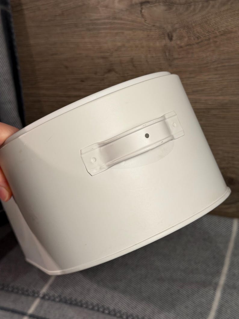 May include: A matte white cylindrical metal container featuring a single horizontal handle attached to the side. The handle is secured with two visible rivets and has a small centered dot detail. The container has a smooth, solid finish.