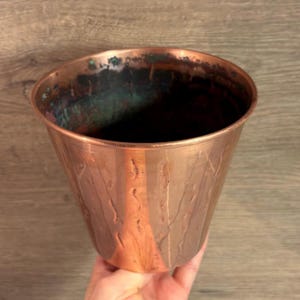 May include: A copper-coloured, cone-shaped planter with a textured surface. The interior shows hints of green oxidation. The planter is held by a hand, showcasing its size and form. Suitable for indoor or outdoor use.