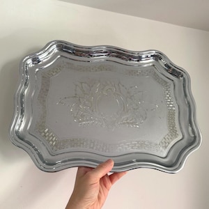 May include: A silver-coloured serving tray with a decorative floral and geometric pattern. The tray has a scalloped edge and a rectangular centre. The tray is made of metal and reflects light.