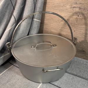May include: A silver pot with a lid and a metal handle. The pot is sitting on a gray and white blanket.