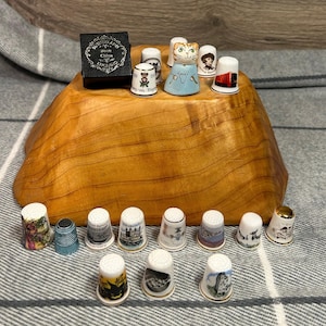 May include: A collection of 20 miniature thimbles, some with a cat, a bus, and a castle design. The thimbles are arranged on a wooden surface. The thimbles are all white with gold trim. The thimbles are all different designs. The thimbles are all in a box.