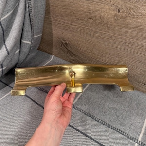 Könnte beinhalten: Ein polierter Kamin-Fender aus Messing mit einem zentralen Knopf. Der Fender hat eine rechteckige Form mit einer geschwungenen Oberseite und einem kleinen, quadratischen Fuß an jedem Ende. Das Messing hat eine helle, reflektierende Oberfläche.