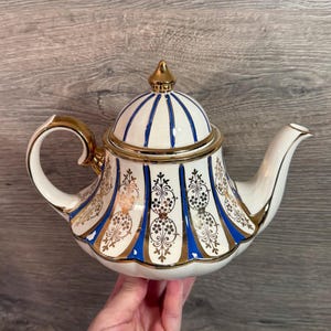 Könnte beinhalten: Eine weiße Keramik-Teekanne mit goldenen Verzierungen und Akzenten, mit einem blau-goldenen Muster. Der Deckel hat eine goldene Spitze und blaue Streifen. Griff und Ausguss sind ebenfalls goldfarben. Ein dekoratives Stück zum Servieren von Tee.