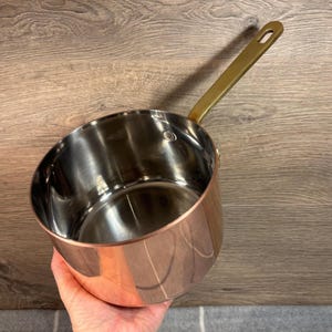 May include: A copper-colored saucepan with a shiny stainless steel interior and a brass-colored handle. The handle is attached to the side of the pan. The pan is held by a person's hand.
