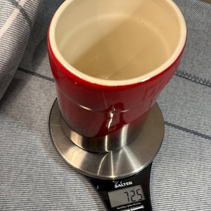 May include: A red and stainless steel kitchen scale with a digital display showing 7.25. The scale is on a gray and white striped surface.