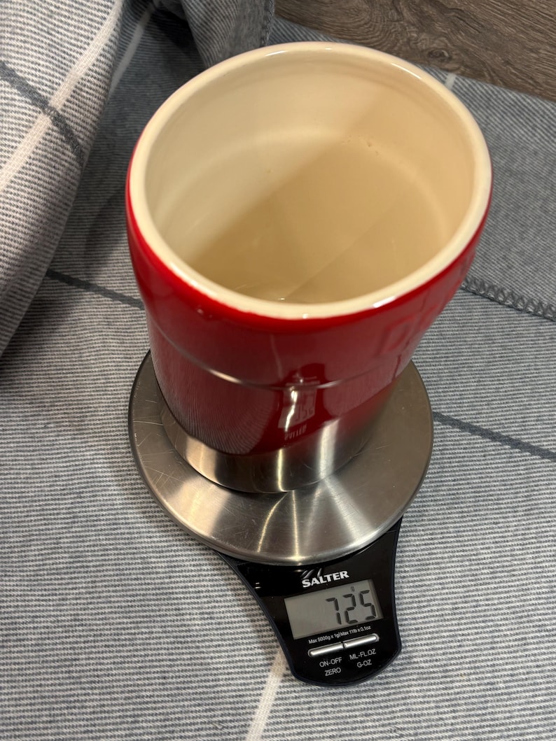 May include: A red and stainless steel kitchen scale with a digital display showing 7.25. The scale is on a gray and white striped surface.