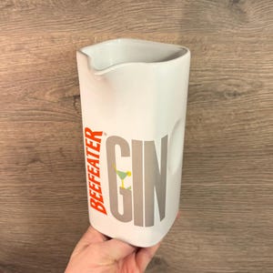 Könnte beinhalten: Weißer Keramik Krug mit einer silbernen und grauen "GIN" Grafik und den Worten "BEEFEATER" in Orange.