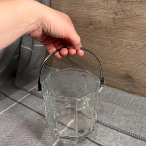 Puede incluir: Hielera de vidrio transparente con forma octogonal y asa de metal plateado. El cubo está vacío y lo sostiene una mano. El fondo es una manta a cuadros gris y blanca y una pared de madera.