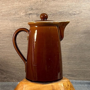 May include: A brown ceramic coffee pot with a lid and a handle. The pot has a glossy finish and a spout for pouring. The lid has a small knob on top. The pot is sitting on a wooden surface.
