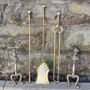 Peut inclure: Un ensemble de cinq accessoires de cheminée en laiton comprenant deux chenets avec des pommeaux en forme de heaume de chevalier, un tisonnier, une pelle à lame en forme de coquille et une pince. Chaque pièce a une finition dorée polie.