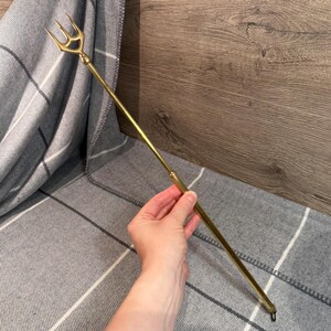 Op de afbeelding: Een goudkleurige drietand met een drietandige kop en een lange, slanke steel. De drietand wordt vastgehouden tegen een grijs-witte geruite deken en een houten oppervlak.