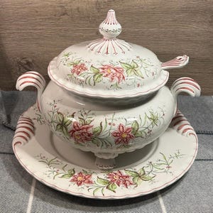 Op de afbeelding: Een witte keramische soepterrine met een deksel, handvatten en een bijpassend bord. De terrine en het bord zijn versierd met roze bloemen- en groene bladerpatronen en rode accenten. De terrine heeft een klassiek ontwerp.