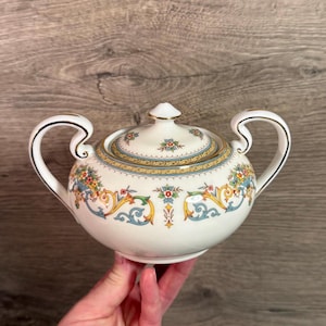 Può includere: Una zuccheriera in ceramica bianca con coperchio, con un motivo floreale e a volute in giallo, blu e rosso. La zuccheriera ha due manici curvi e un bordo dorato. Il coperchio è sormontato da un piccolo pomello.