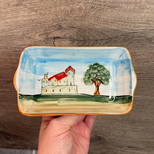 Peut inclure: Plat de service rectangulaire en céramique avec une scène de paysage peinte à la main. Le motif représente un bâtiment blanc avec un toit rouge, un arbre vert et un ciel bleu. Le plat a un bord et des poignées oranges.