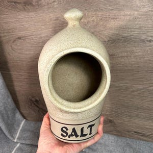 Puede incluir: Una salera de cerámica con un esmalte beige moteado y una inscripción "SALT" en negro. La salera tiene una abertura redonda en la parte superior para verter sal.