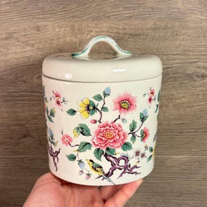 Pode incluir: Um pote de cerâmica branco com um design floral com flores rosa, amarelas e azuis e um pequeno pássaro amarelo. O pote tem uma tampa com uma alça verde.