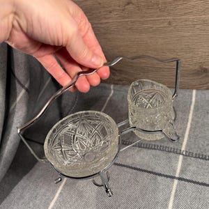 May include: A vintage clear glass serving set with a decorative cut-glass pattern. The set includes two bowls and a handle, all held together by a silver-colored metal frame. The bowls are round and sit on small feet.